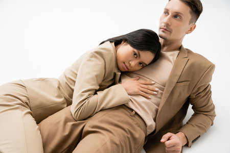 sensual asian woman looking at camera while hugging trendy man lying on white backgroundの写真素材