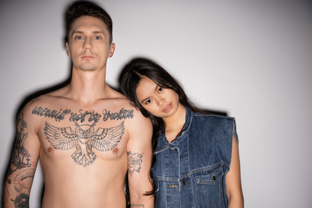 front view of shirtless tattooed man and asian woman in denim vest looking at camera on grey backgroundの写真素材