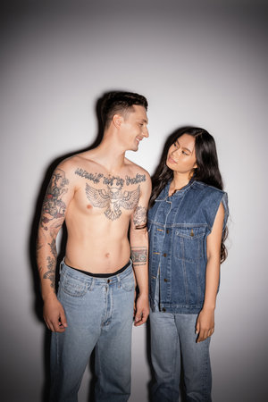 shirtless tattooed man and asian woman in denim clothes smiling at each other on grey backgroundの写真素材