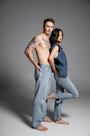full length of tattooed shirtless man looking at camera near stylish asian woman in denim clothes on grey backgroundの写真素材