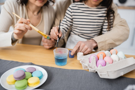Cropped view of family holding paintbrushes near paints and Easter eggs in kitchenの写真素材