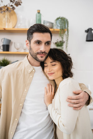 Bearded man hugging smiling wife in kitchen at homeの写真素材