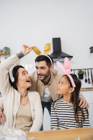 Smiling woman looking at Easter headband on husband near daughter at homeの写真素材