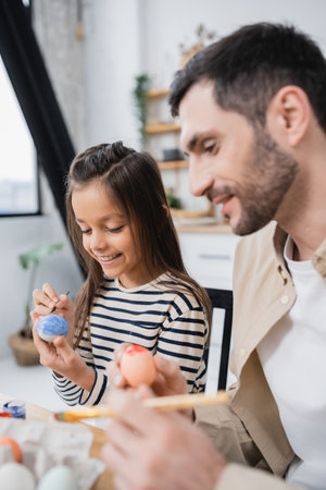Smiling girl painting Easter eggs near blurred father at homeの写真素材
