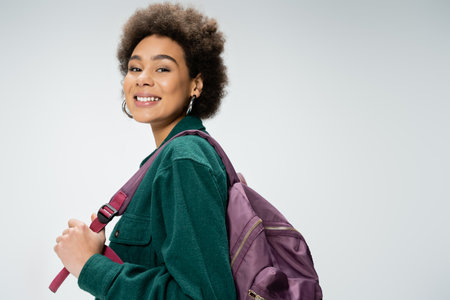cheerful african american woman in hoop earrings posing with backpack isolated on greyの写真素材