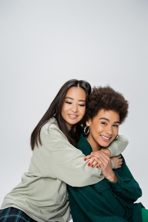 brunette asian woman hugging cheerful african american friend looking at camera isolated on greyの写真素材
