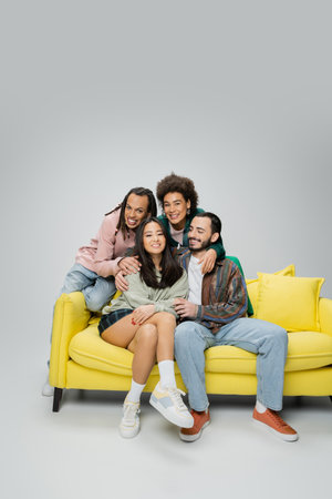smiling multiracial friends in fashionable clothes posing near yellow sofa on grey backgroundの写真素材