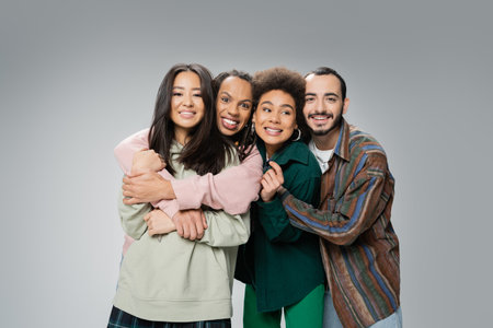 joyful and fashionable multicultural friends posing and hugging isolated on greyの写真素材