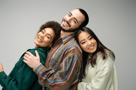 joyful multicultural friends in trendy outfit smiling at camera while posing isolated on greyの写真素材