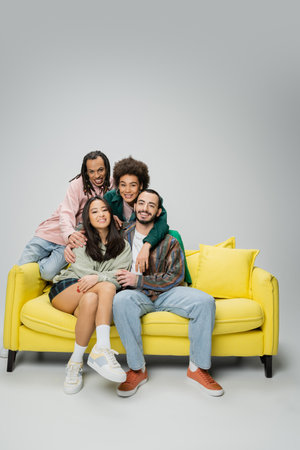 happy and stylish multicultural friends posing near yellow couch and looking at camera on grey backgroundの写真素材