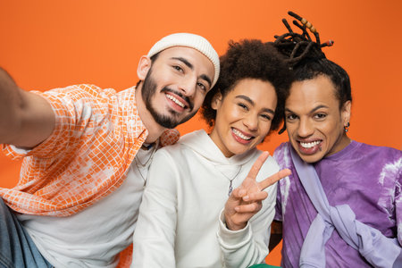 joyful african american woman showing victory sign near trendy multiethnic friends isolated on orangeの写真素材