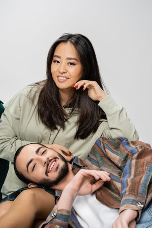 happy interracial couple in trendy clothes smiling at camera isolated on greyの写真素材