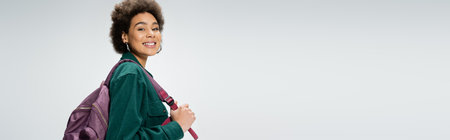 pleased and stylish african american student with backpack looking at camera isolated on grey,...