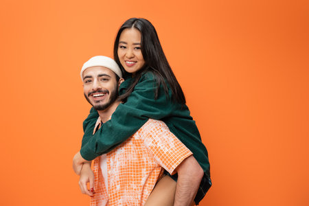 young bearded man in beanie piggybacking cheerful asian woman isolated on orangeの写真素材