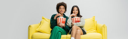 young and cheerful interracial women eating popcorn and watching movie on yellow couch isolated on grey, bannerの写真素材