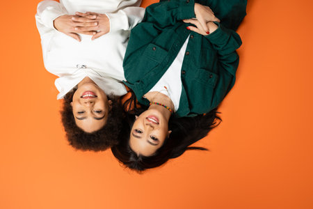 top view of happy african american and asian women in stylish attire lying on orange backgroundの写真素材