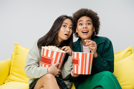 amazed interracial women holding buckets of popcorn while watching movie on yellow couch isolated on greyの写真素材