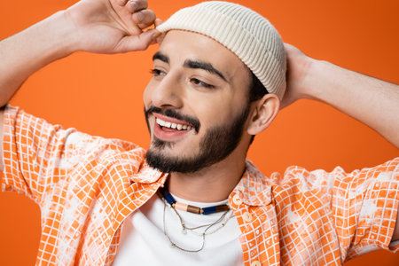 portrait of cheerful bearded man in beads touching white beanie and looking away isolated on orangeの写真素材
