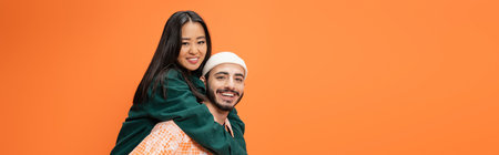 happy bearded man in beanie piggybacking pretty asian woman isolated on orange, banner