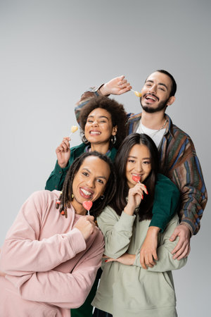 happy multiethnic friends in stylish outfit posing with sweet lollipops isolated on greyの写真素材