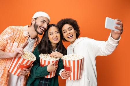 happy african american woman taking selfie with trendy friends holding buckets of popcorn isolated on orangeの写真素材