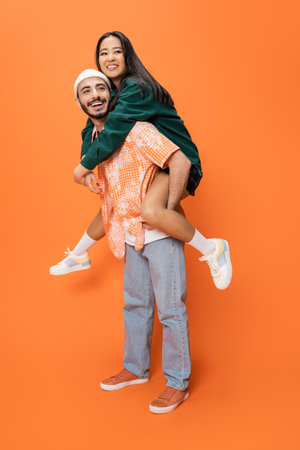 full length of excited man in beanie and jeans piggybacking trendy asian woman on orange backgroundの写真素材