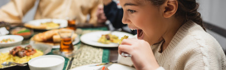 Muslim girl eating iftar dinner near blurred food and parents at home, bannerの写真素材