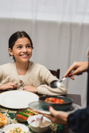 Smiling muslim girl looking at blurred mom with food during ramadan dinner at homeの写真素材