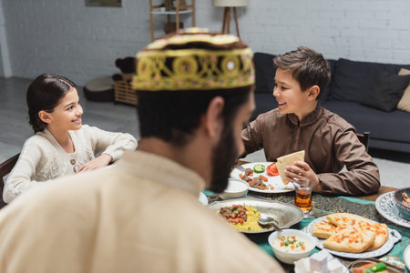 Positive muslim kids sitting near food and blurred father during ramadan at homeの写真素材
