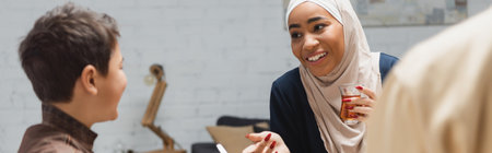 African american mother in hijab holding tea and talking to son at home, bannerの写真素材