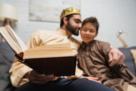Book in hand of muslim man hugging son on couch at homeの写真素材