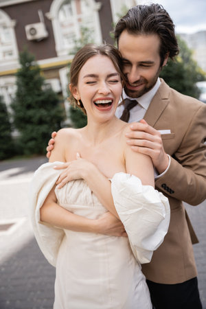 portrait of cheerful and bearded man hugging happy bride laughing with closed eyesの写真素材