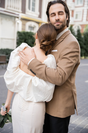 elegant and bearded groom with closed eyes hugging bride in white dressの写真素材