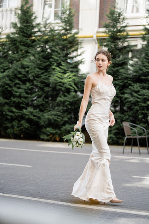 full length of young woman in white dress holding wedding bouquet of blooming flowers and standing outsideの写真素材
