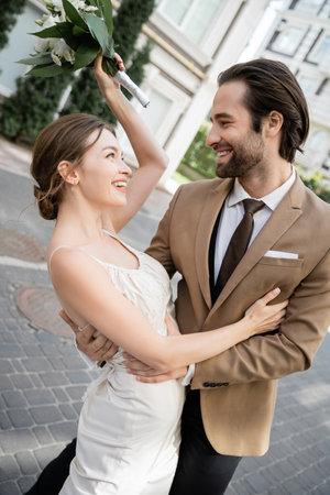 joyful bride in wedding dress holding bouquet and smiling with groomの写真素材