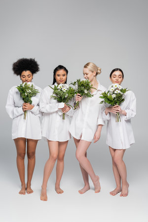 full length of young women with bare legs wearing white shirts and enjoying flavor of aromatic flowers on grey backgroundの写真素材