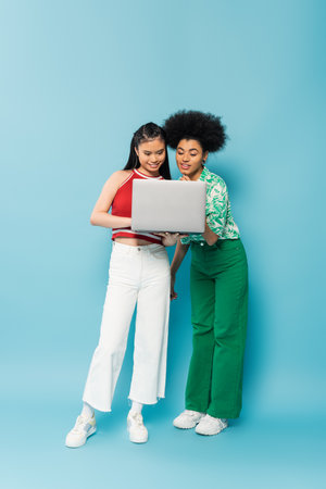 full length of young multiethnic woman in trendy clothes smiling while looking at laptop on blue backgroundの写真素材
