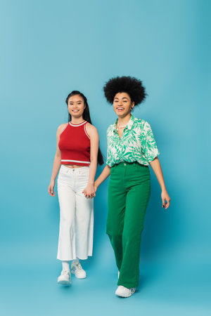 full length of joyful multiethnic women in trendy clothes holding hands while walking on blue backgroundの写真素材