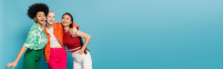 joyful multicultural women in trendy and colorful clothes embracing and smiling at camera isolated...