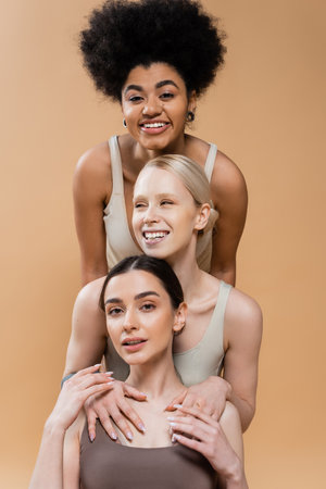 pretty and cheerful multiethnic models in lingerie posing and smiling isolated on beigeの写真素材