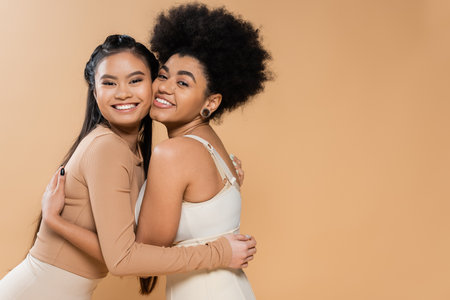 joyful asian and african american women in underwear embracing and looking at camera isolated on beigeの写真素材