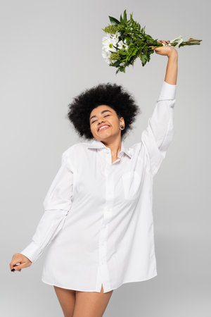 young african american woman in white shirt holding flowers in raised hand and smiling with closed eyes isolated on greyの写真素材