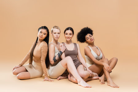 full length of young multiethnic women in underwear sitting and looking at camera on beige backgroundの写真素材