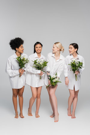 full length of barefoot multicultural women in white shirts holding bouquets on grey backgroundの写真素材
