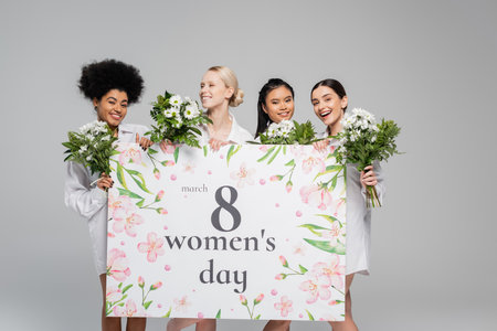 young interracial women smiling near flowers and huge womens day greeting card isolated on grey