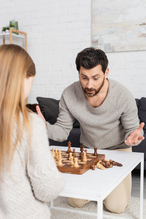 confused bearded man showing shrug gesture while playing chess with preteen daughter at home