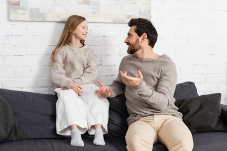 bearded man gesturing while talking to smiling daughter on couch in living roomの写真素材