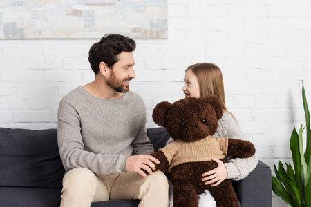 happy girl holding soft toy and looking at cheerful bearded dad while sitting on sofa at homeの写真素材
