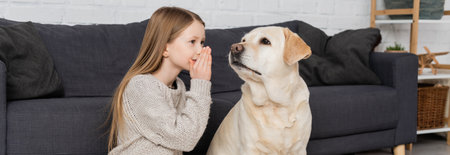 preteen girl covering mouth with hand while telling secret to labrador dog at home, bannerの写真素材