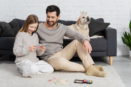 happy father and daughter looking at makeup palette while sitting near labrador dog in living roomの写真素材